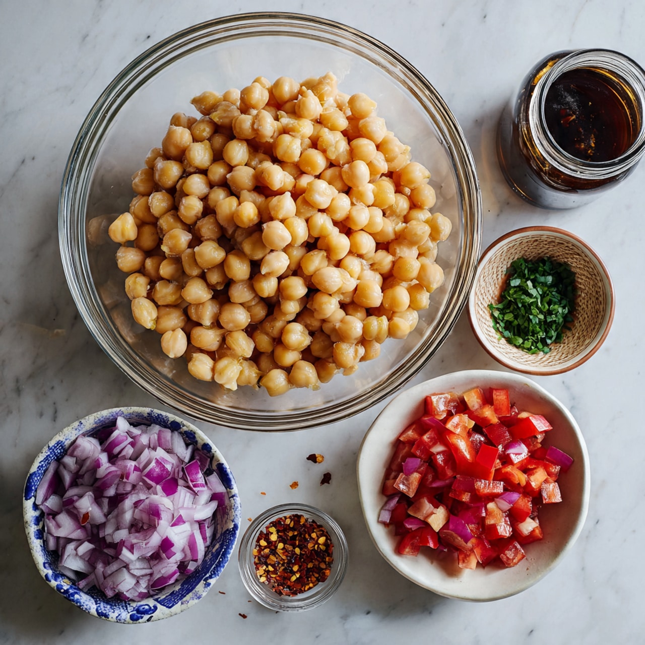 The image shows a clear glass bowl filled with light yellow chickpeas as the main central layer, resting on a white marbled surface. Surrounding the bowl are smaller white bowls with blue patterns containing diced red onions and diced red tomatoes, adding purple and red colors. There are also small dark bowls holding chopped green herbs and a glass jar of dark brown sauce. Additionally, a small glass spice jar with reddish chili flakes is placed near the center. The overall arrangement of the ingredients is neat and colorful, set on the white marbled surface with a soft natural light, photo taken with an iphone --ar 4:5 --v 7