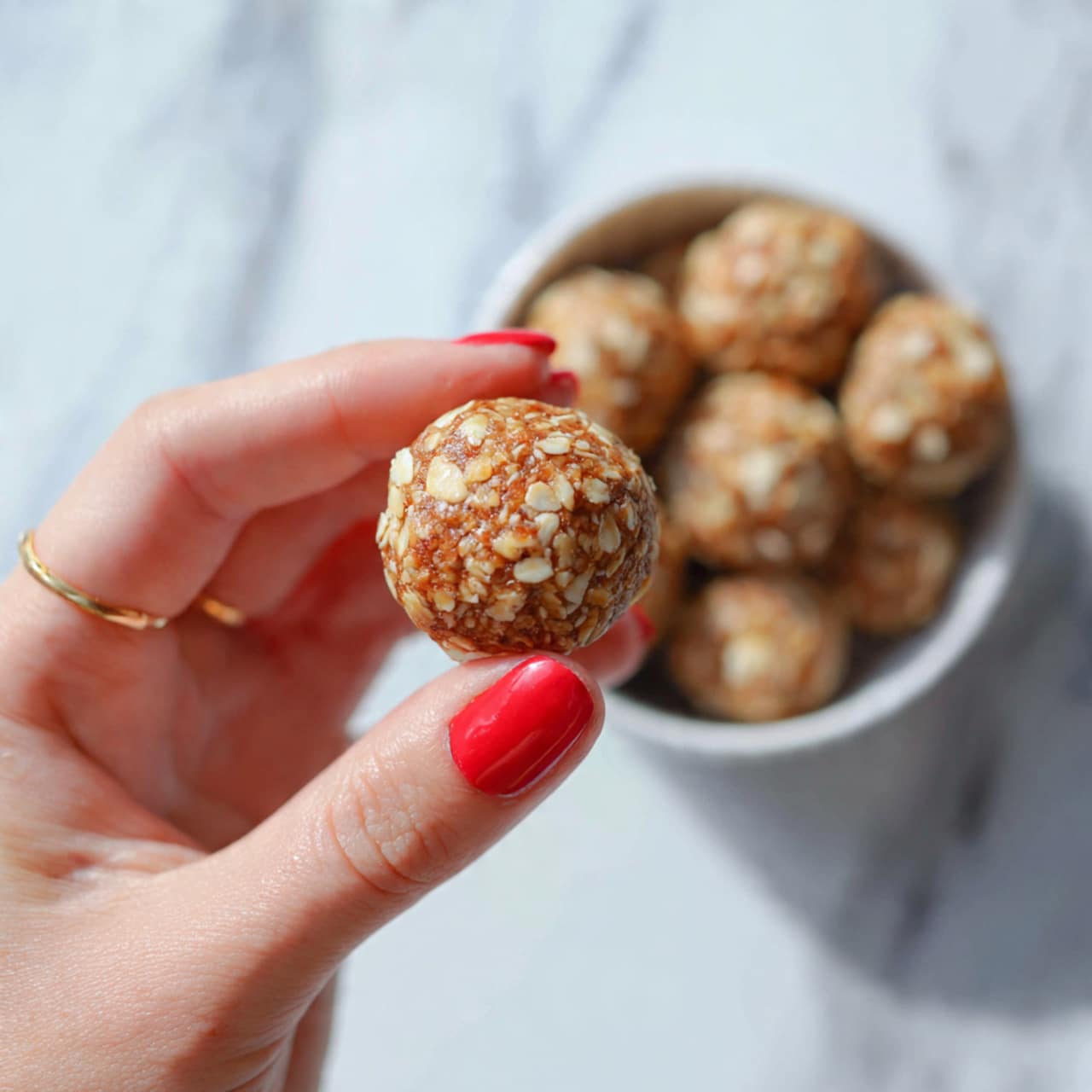 Two light brown energy balls with small white nut pieces are stacked on a white marbled surface; the top ball is bitten to show a rough, crumbly inside texture, while three more blurred energy balls appear in the background, softly out of focus photo taken with an iphone --ar 4:5 --v 7