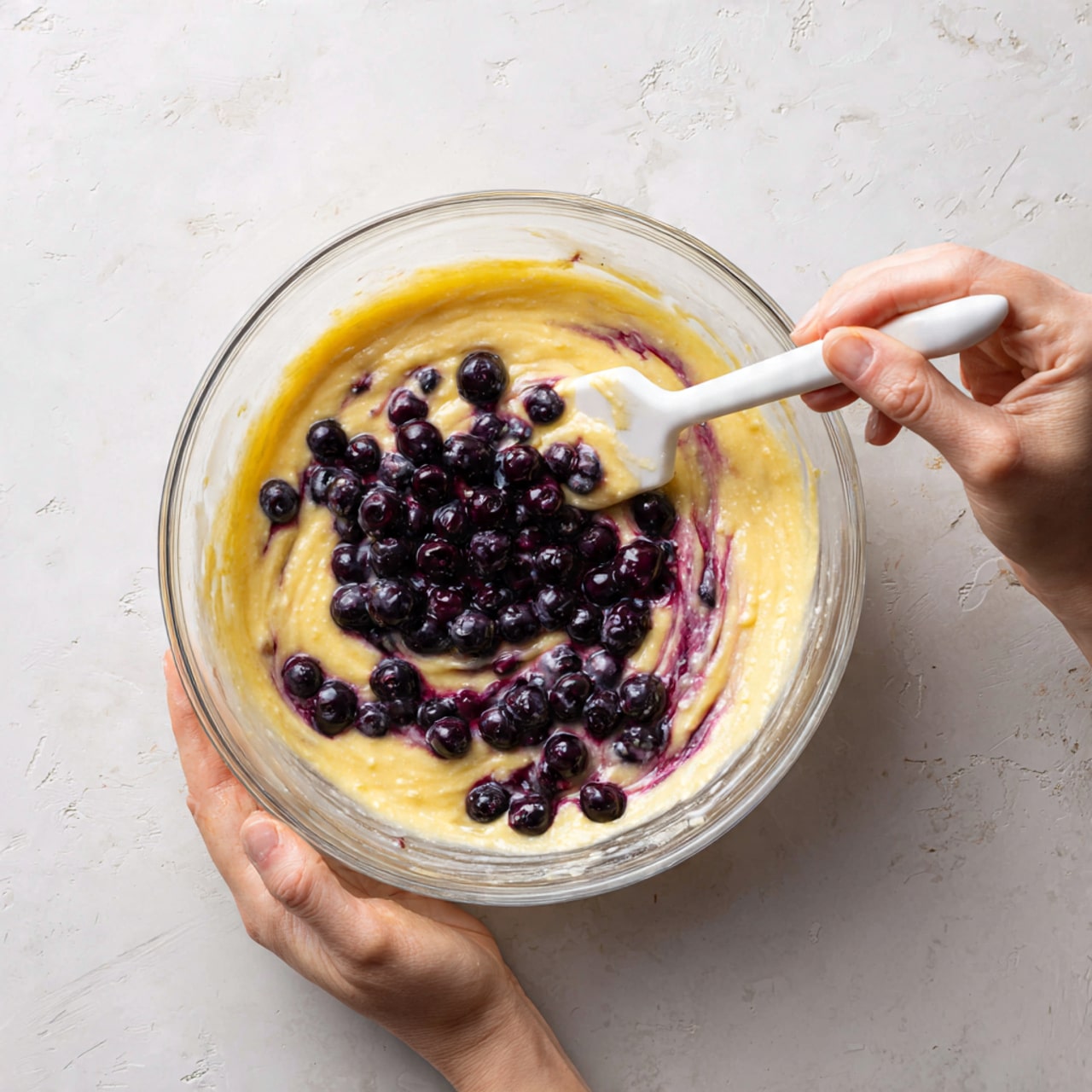 A clear glass bowl contains a creamy yellow batter mixed with whole dark blueberries. The batter shows swirls of deep purple from the blueberry juice blending into it. The blueberries are mostly on the surface but some are partly submerged. A white spatula is lifting some of the batter and berries from the bowl. A woman's hand is gently holding the side of the bowl, which sits on a white marbled textured surface. The texture looks smooth and slightly thick, with the blueberries adding contrast. Photo taken with an iphone --ar 4:5 --v 7