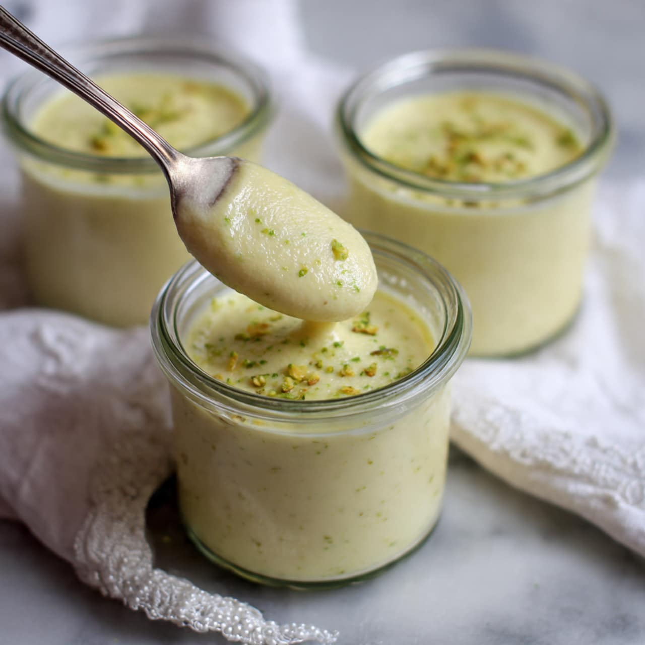 The image shows three small glass jars filled with a smooth, creamy yellow pudding. Each jar is topped with a sprinkle of green and white chopped nuts, adding texture and color contrast. Two jars in the background have silver spoons inside, while the one in the front does not. The jars are placed on soft white fabric with delicate embroidery, all set against a white marbled surface. The overall look is soft, light, and inviting, highlighting the creamy dessert and the natural tones of the nuts photo taken with an iphone --ar 4:5 --v 7