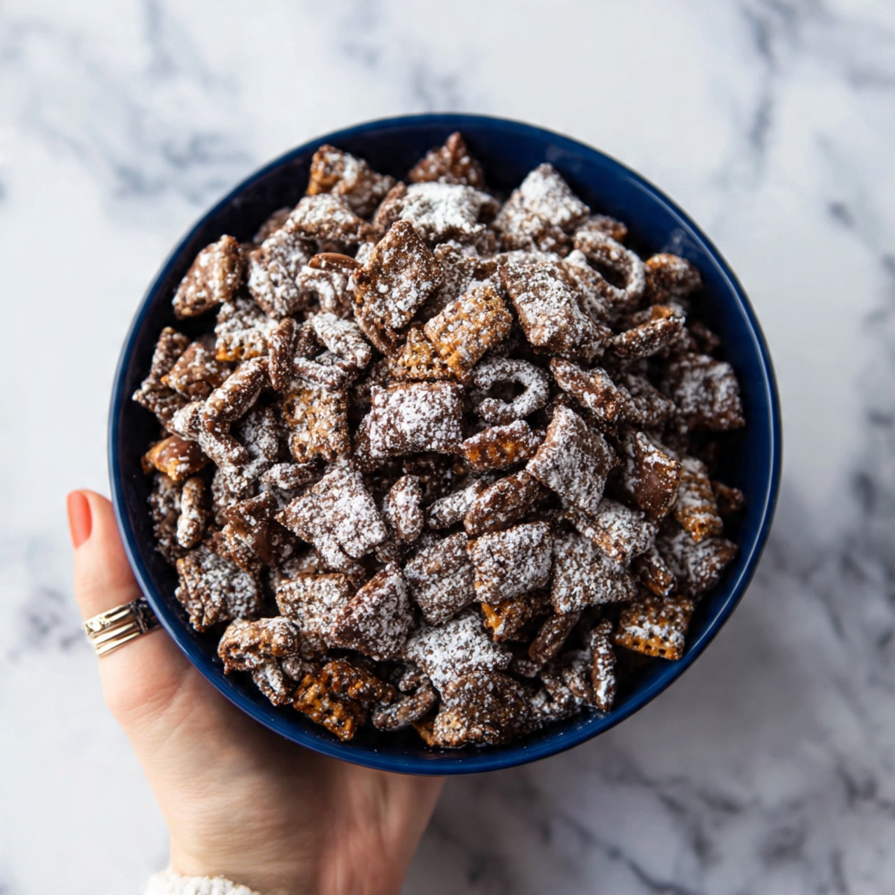 A dark blue bowl full of chocolate and powdered sugar coated snack mix, showing a mix of small and medium bite-sized crunchy pieces with rough and uneven textures piled high inside. A woman's hand with a silver ring at the base of her finger is holding the bottom edge of the bowl, positioned in the lower right corner of the image. The bowl sits on a white marbled surface with light grey veins running through it. The overall scene is bright and detailed, highlighting the sugary coating and textured layers of the snack mix. photo taken with an iphone --ar 4:5 --v 7