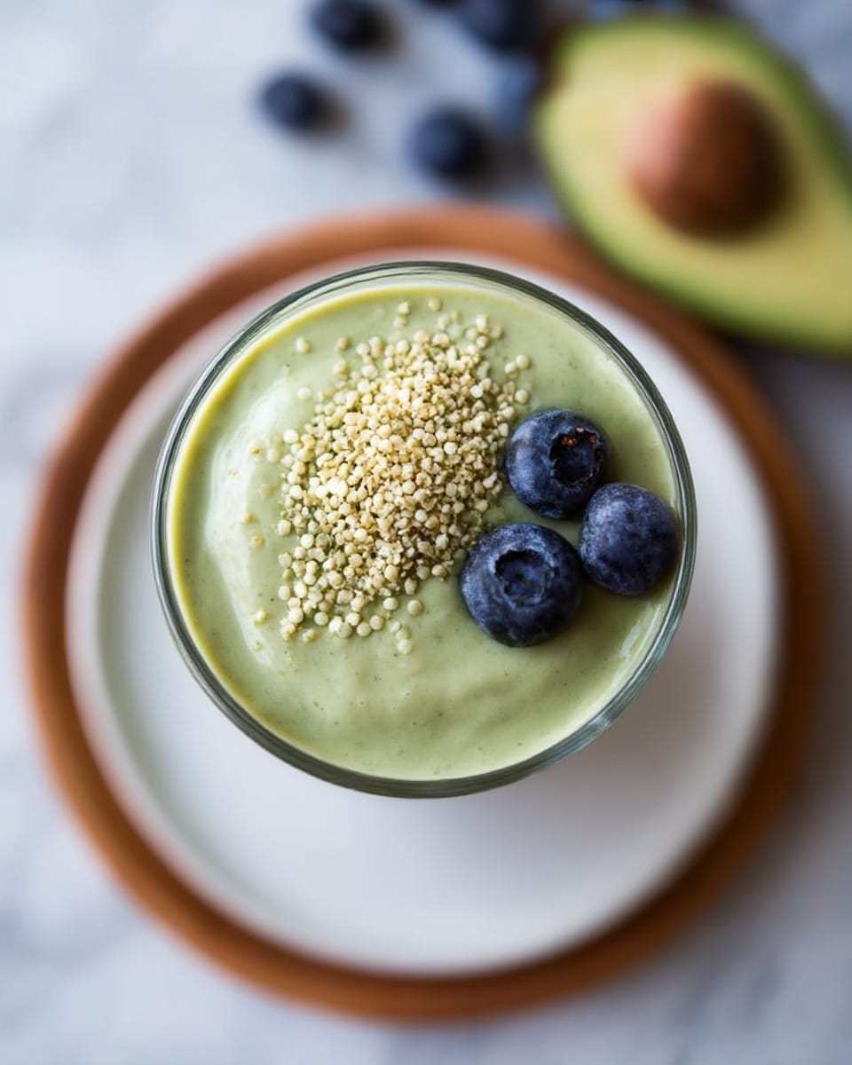 A tall clear glass is filled with a thick light green smoothie with a creamy texture. On top, there are a few fresh blueberries clustered in the center and a small pile of light beige hemp seeds to the right side. The glass sits on a round wooden plate with more blueberries scattered loosely around its base. In the background, there is a halved avocado with a light green flesh and a brown seed visible. The setting is on a white marbled surface. Photo taken with an iphone --ar 4:5 --v 7