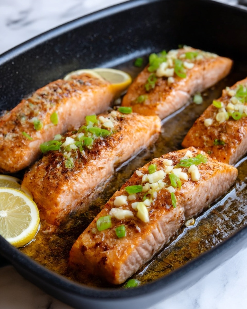 Four cooked salmon fillets with a golden brown crust and tender pink inside lie in a black pan, each topped with small bits of white garlic and chopped green onions. Thin slices of light yellow lemon rest under and beside the fish, adding brightness. The pan has a slightly shiny surface with some cooking oil visible, creating a rich texture. The whole scene is on a white marbled surface. photo taken with an iphone --ar 4:5 --v 7