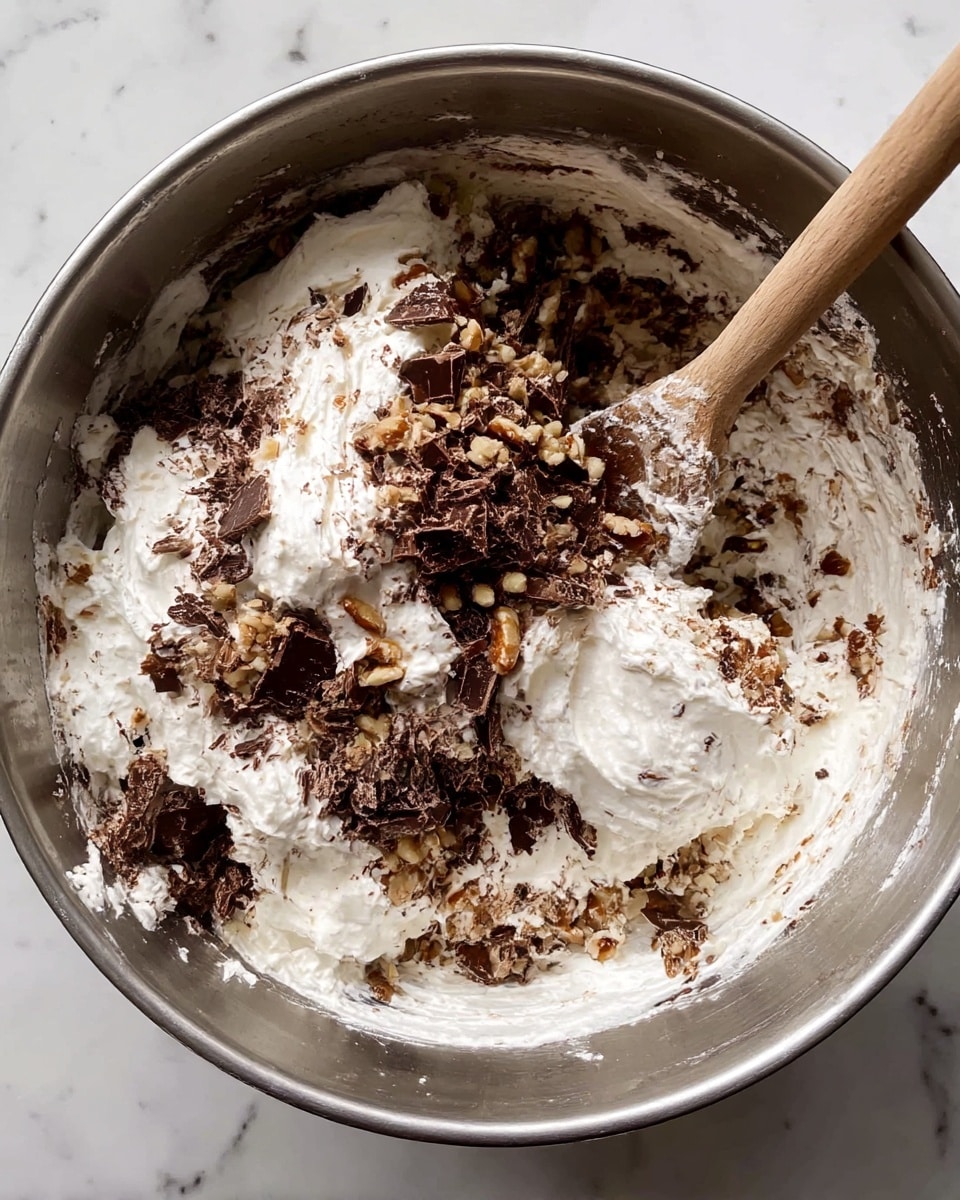 Inside a silver metal bowl, there is a mix of different ingredients. One layer is fluffy white cream that looks soft and thick. Mixed with it are chopped dark brown chocolate pieces, adding texture and color. There are also small pieces of light brown nuts sprinkled over the cream and chocolate, giving a rough and crunchy look. A wooden spoon is partly stuck into the mix, with some cream and chocolate on it, showing the mixing process. The background under the bowl is a white marbled texture. photo taken with an iphone --ar 4:5 --v 7
