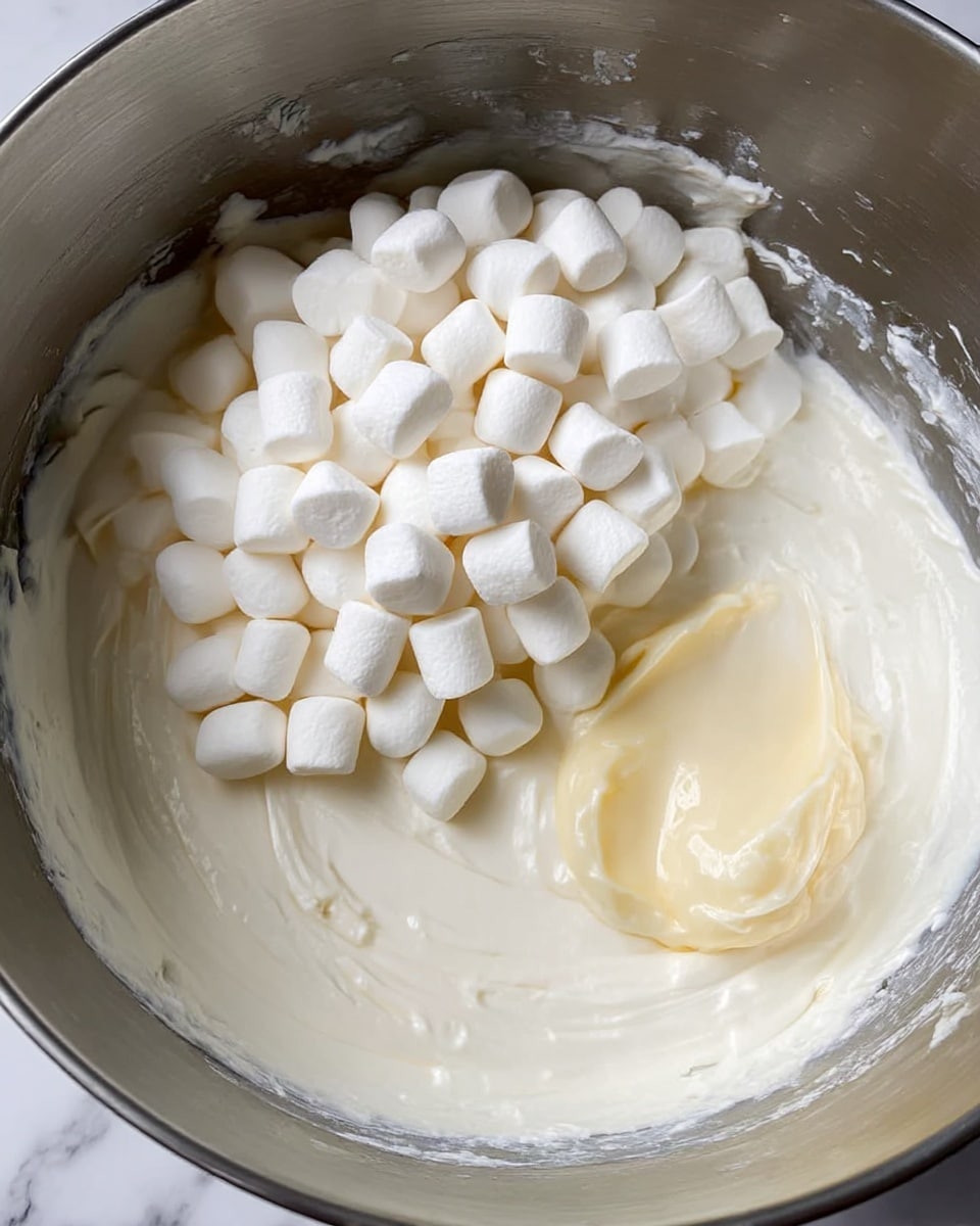 A close-up shot of a metal mixing bowl filled halfway with a smooth, thick white cream base. On top of the cream, there is a pile of small, white mini marshmallows. To the right of the marshmallows, there is a dollop of a pale yellow substance, slightly glossy and smooth. The inside walls of the bowl have some cream residue, adding texture. The background is a white marbled surface. Photo taken with an iphone --ar 4:5 --v 7