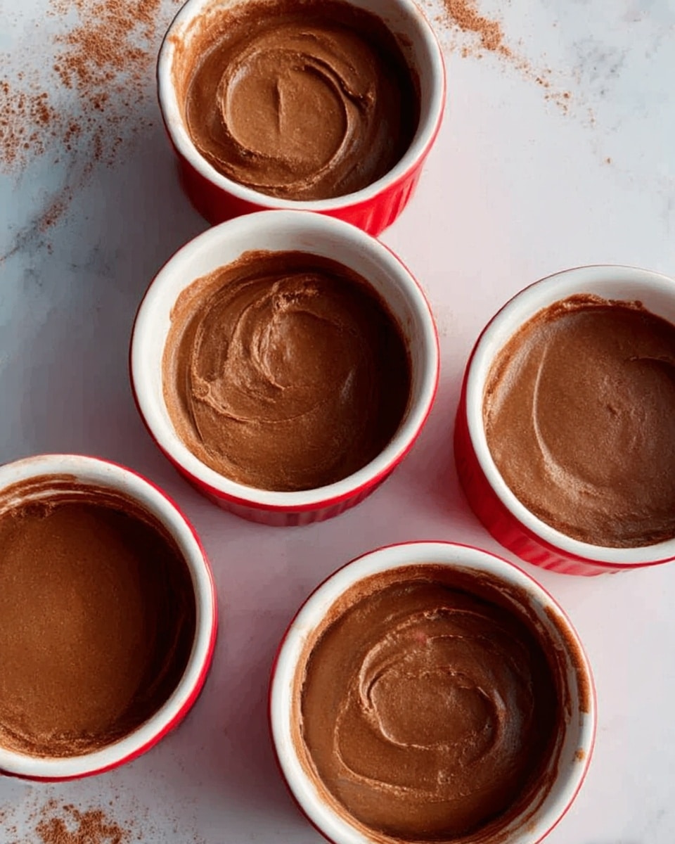 Four red ramekins with white rims are shown from above, each filled with a smooth, even layer of creamy chocolate pudding that coats the inside surfaces. The pudding has a glossy dark brown color with a slightly swirled texture from spreading. The ramekins are placed on a white marbled surface with some faint brown powder scattered lightly around them. The photo taken with an iphone --ar 4:5 --v 7
