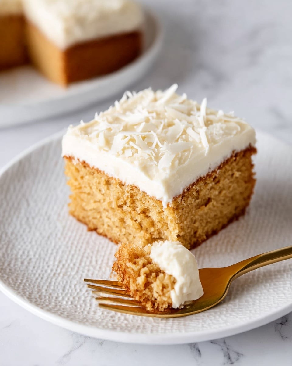 A square piece of golden-brown cake with a thick layer of creamy off-white frosting on top, sprinkled with white chocolate shavings. The cake is placed on a white plate with a textured surface. In front of the cake, a gold fork holds a small bite-sized piece of cake that shows the soft crumb and frosting. The background is a white marbled texture. Photo taken with an iphone --ar 4:5 --v 7