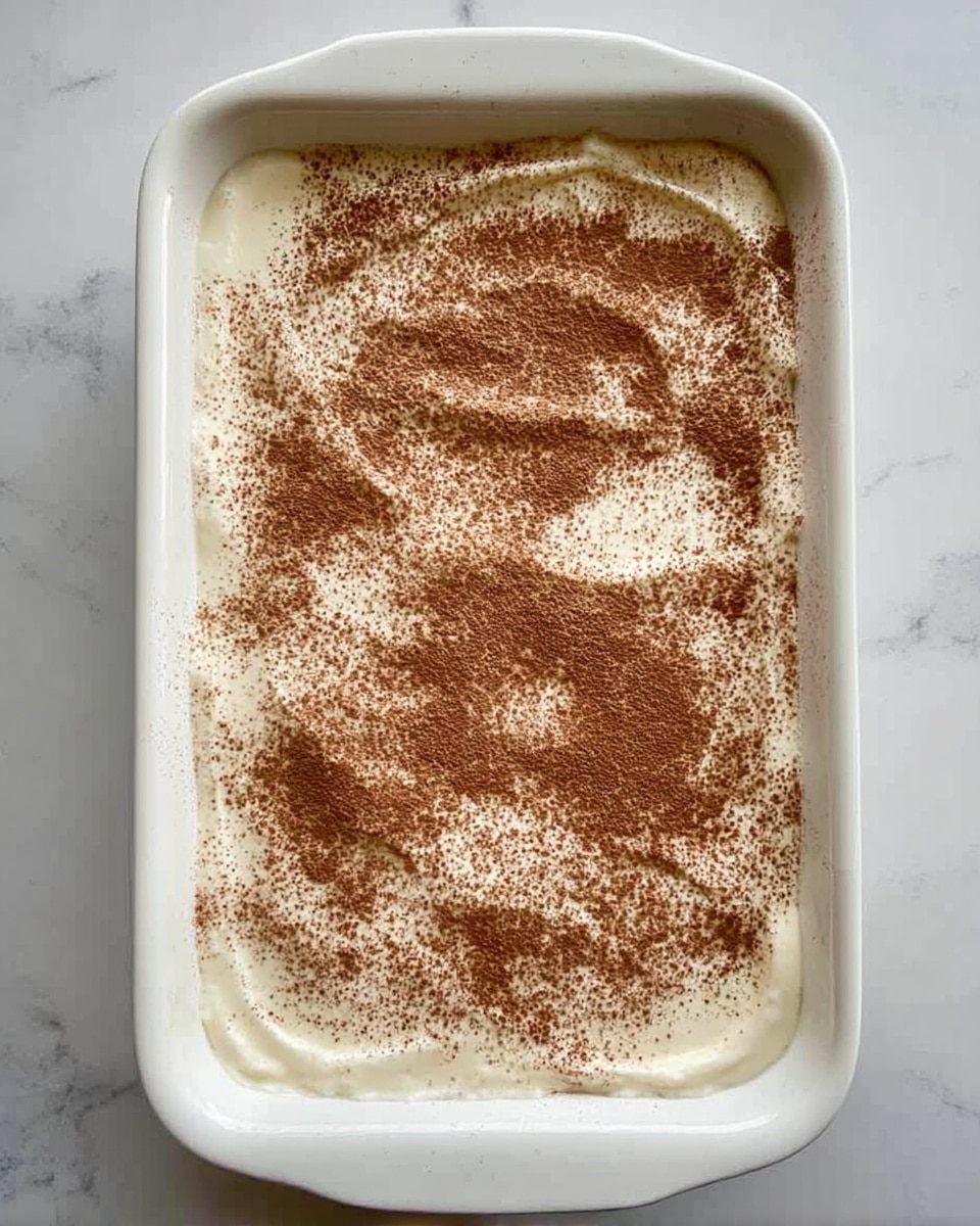 A white rectangular baking dish filled with one layer of smooth, creamy white dessert, dusted unevenly with a light brown powder on top. The powder is scattered in a natural, random pattern across the surface, covering most but not all the cream beneath. The dish is placed on a white marbled surface, with the edges of the dish clearly visible and clean. photo taken with an iphone --ar 4:5 --v 7