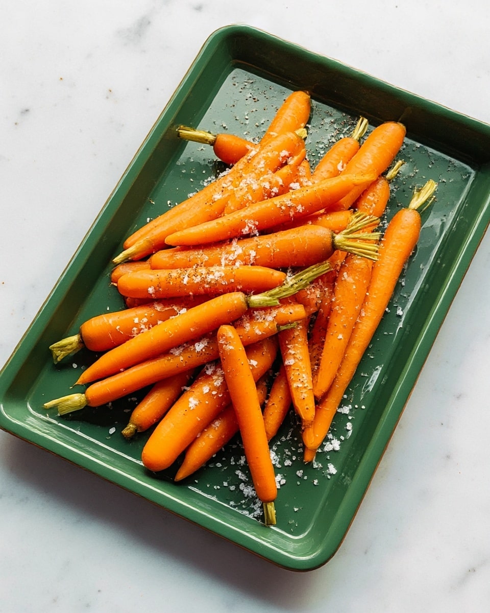 A green rectangular tray holds about twenty small orange carrots scattered over it. The carrots are smooth and fresh with green tops still attached at some ends. On top of the carrots and tray are coarse sea salt grains and crushed black pepper sprinkled unevenly, adding texture and contrast to the bright carrots. The tray rests on a white marbled surface. photo taken with an iphone --ar 4:5 --v 7