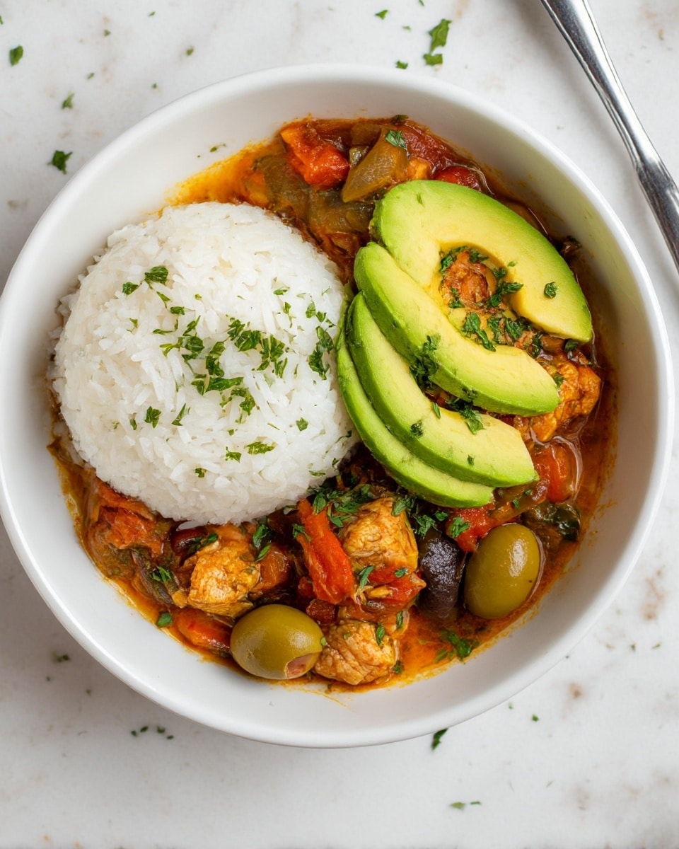 The dish shows a white bowl filled with two main layers. One layer is a round mound of white rice on the left side, fluffy and slightly sprinkled with green herbs. The second layer covers the right side and is a stew with orange and brown tones, containing pieces of cooked chicken, green olives, sliced onions, and green and red bell pepper slices. On top of the rice layer are three thin slices of bright green avocado, fanned out. Small bits of chopped green herbs scatter over the stew and around the bowl on a white marbled surface. Nearby are parts of another similar bowl, a wooden bowl with coarse white salt, and a small bowl with chopped green herbs. Photo taken with an iphone --ar 4:5 --v 7