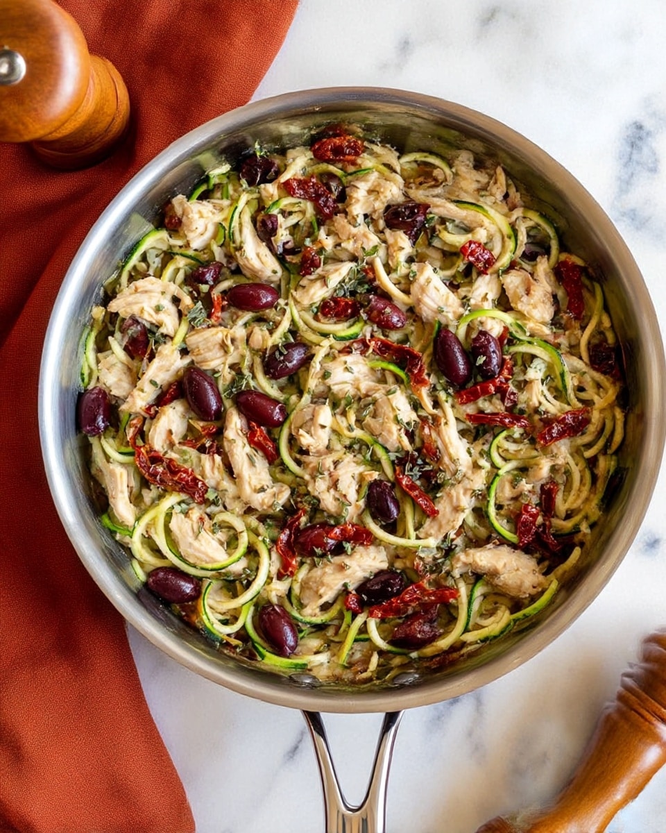 A shiny metal pan filled with layers of light brown cooked chicken pieces scattered on top of thin green spiralized zucchini noodles, mixed with dark purple olives and bright red sun-dried tomato strips. Small green herb flakes are sprinkled over the dish, adding texture and color contrast. The pan handle is visible at the bottom, placed on a white marbled surface next to a folded rust-colored cloth and a wooden pepper grinder. The overall look is fresh and colorful, with varied textures from the juicy chicken, soft zucchini, and chewy olives. photo taken with an iphone --ar 4:5 --v 7