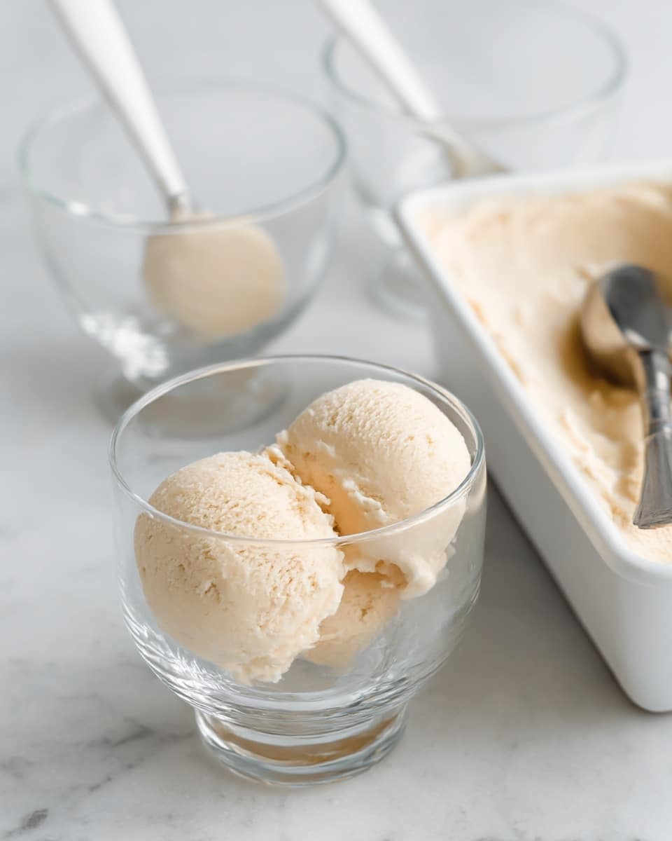The image shows a clear glass bowl with three scoops of light beige ice cream inside it. A metal spoon is placed in the bowl, leaning to the left, with some ice cream touching the inside of the bowl near the bottom. In the background, there are two more empty clear glass bowls with spoons inside them and another bowl on the right containing more light beige ice cream, all set on a white marbled surface. The overall scene is soft and bright with a simple look. photo taken with an iphone --ar 4:5 --v 7