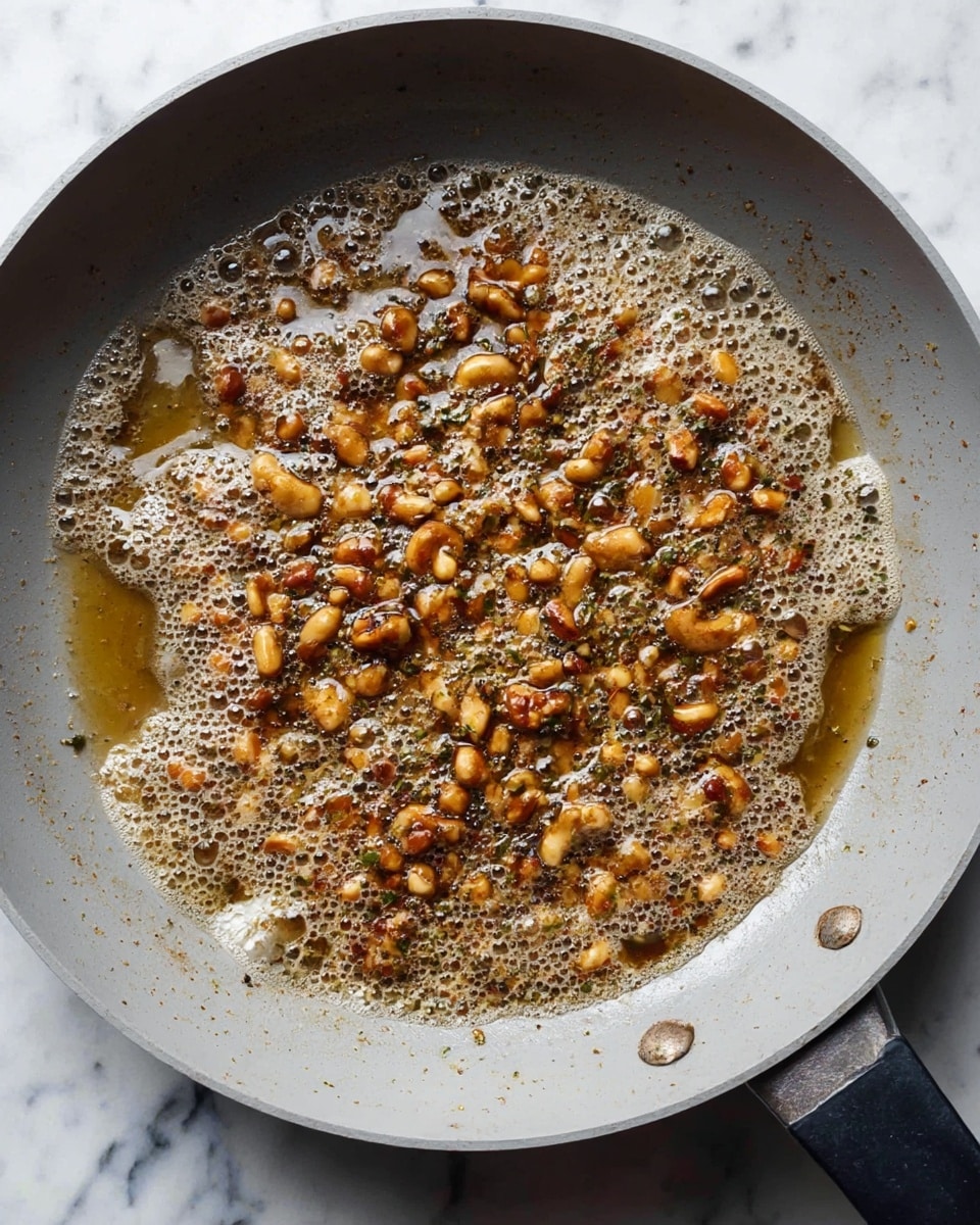 A close-up view of a light gray pan filled with a bubbling, frothy mixture of browned chopped nuts and herbs, spreading unevenly across the pan's surface. The nuts have a golden to deep brown color with a slightly crispy texture, mixed with small green herb bits and light-rich bubbles around them. The pan is placed on a white marbled surface, with part of the pan’s black handle visible at the bottom right corner. The scene shows the cooking process with textures blending in a glossy, oily layer. photo taken with an iphone --ar 4:5 --v 7