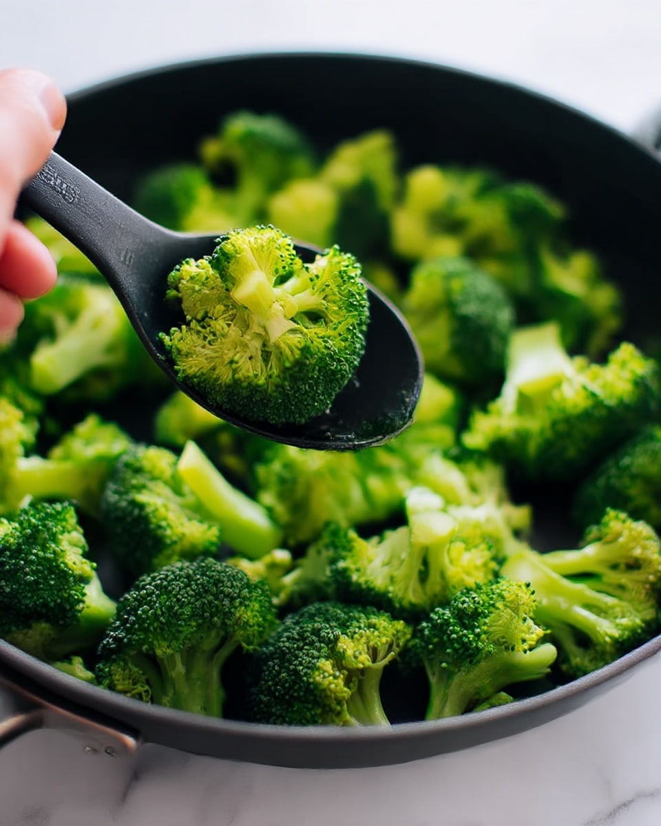 The image shows bright green broccoli florets with small tree-like shapes and bumpy textures inside a black pan. The broccoli pieces are fresh and cooked lightly, giving a mix of darker and lighter green colors. There is a black spoon in the pan lifting some broccoli, and a blurred woman's hand is holding the spoon on a white marbled surface. The focus is sharp on the broccoli, with soft light emphasizing its fresh color. photo taken with an iphone --ar 4:5 --v 7