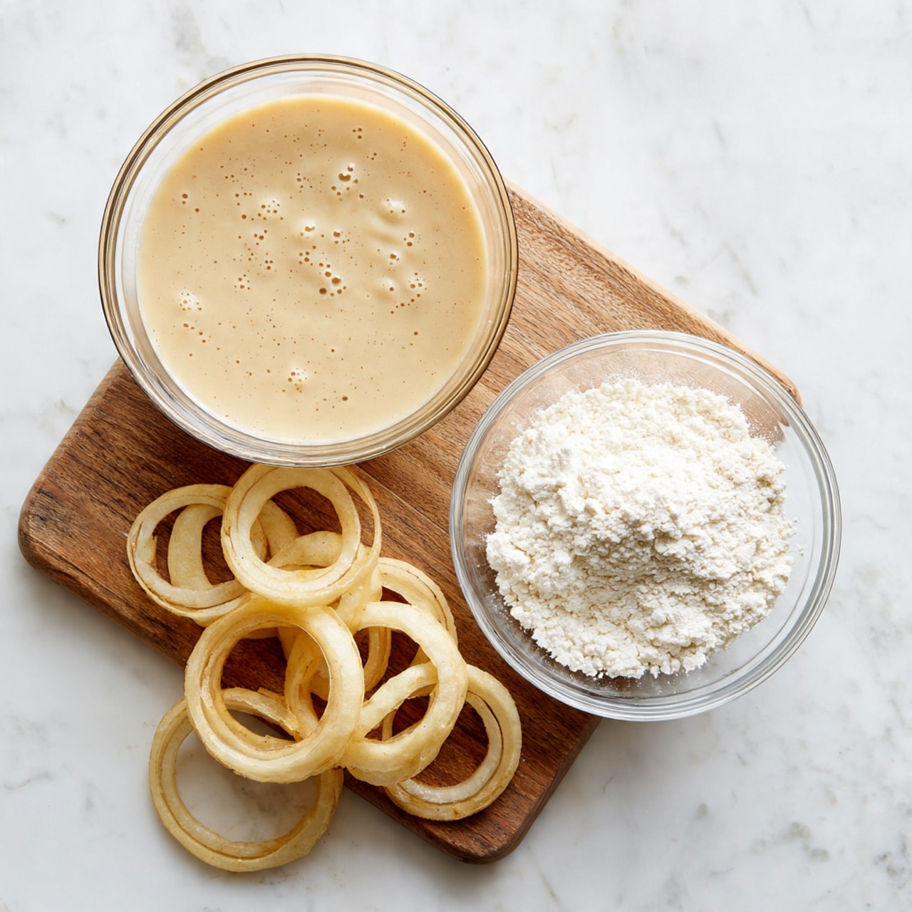 On a white marbled surface, there is a wooden board with thick sliced onion rounds and some separated onion rings scattered on it. Next to the board, there are two clear glass bowls; the top one contains a smooth beige batter with small bubbles on the surface, and the bottom one has white flour dusted inside. The setup shows all ingredients ready for making onion rings or similar fried onion dish. photo taken with an iphone --ar 4:5 --v 7