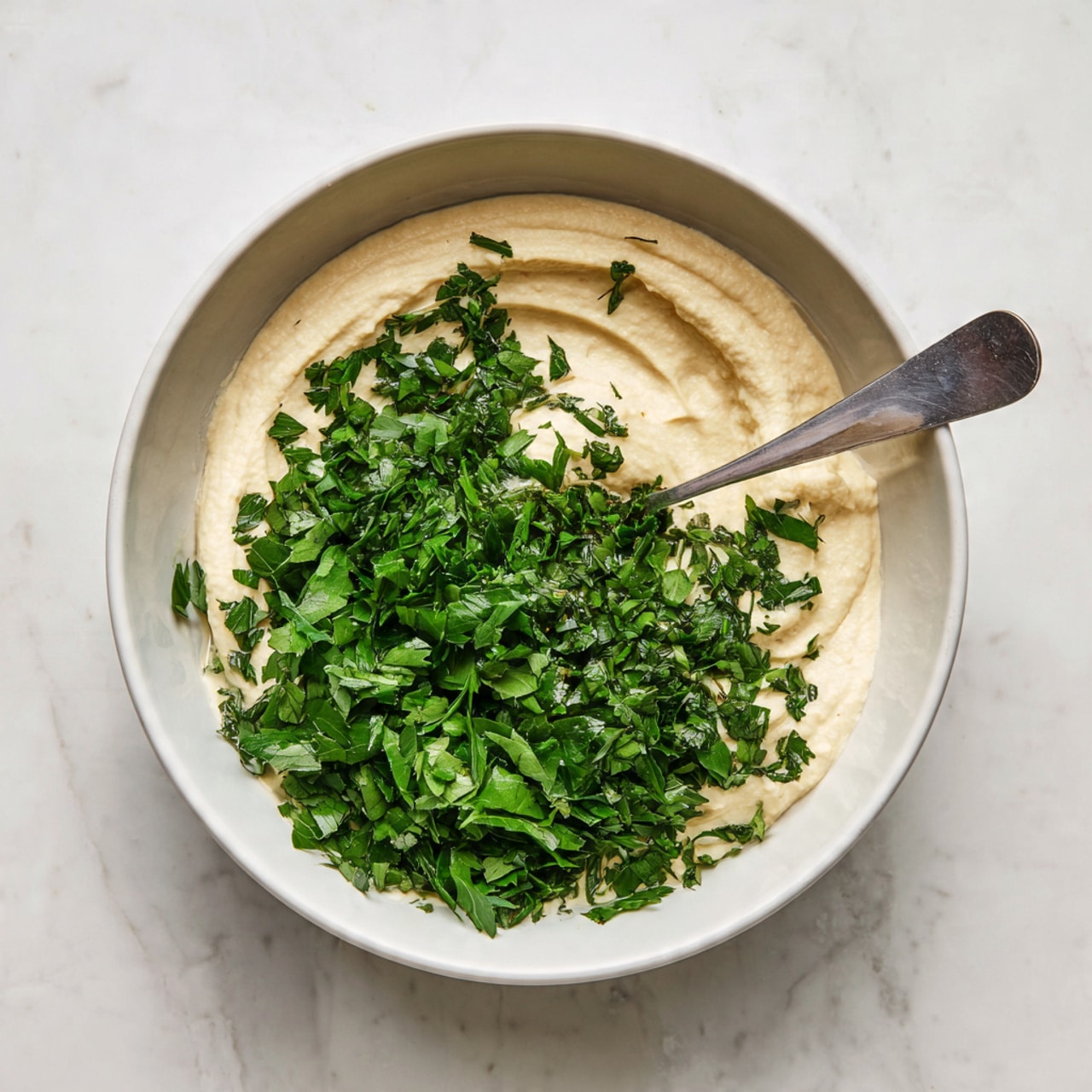 A white bowl filled with two main layers: the bottom layer is a thick, creamy beige sauce, and the top layer is a generous amount of finely chopped fresh green herbs spread unevenly around the sauce. A shiny silver spoon rests inside the bowl, dipping into the green herbs on the right side. The bowl is placed on a white marbled surface with a soft pattern of veins and smooth texture. photo taken with an iphone --ar 4:5 --v 7
