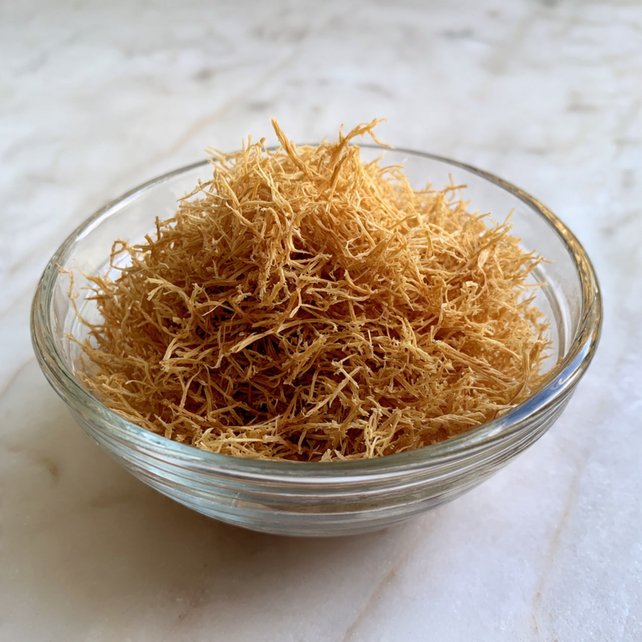 A clear glass bowl filled to the top with a large mound of fine, thin, golden-brown shredded strands that look light and fluffy, resting on a white marbled surface. The texture of the shreds appears delicate and slightly dry, filling the bowl evenly without any visible layers or variation in color. Photo taken with an iphone --ar 4:5 --v 7