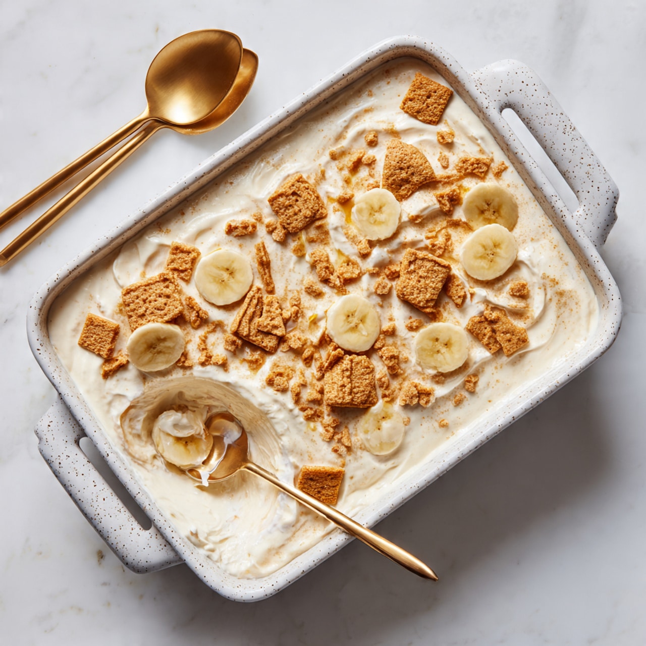 The dish is in a white, speckled, rectangular baking dish with handles on both sides, set on a white marbled surface. It has a creamy white base with smooth texture, spread evenly in two main visible layers. On top, there are irregularly placed broken cookie pieces of light brown color and round slices of banana scattered across, showing soft yellow and white tones with a slightly moist texture. Two gold spoons rest on the dish’s left lower side, slightly scooping into the creamy base, revealing some of the inner layers beneath. The overall look is soft and inviting with visible contrast between the creamy base and the crunchy cookie pieces and fresh banana slices. photo taken with an iphone --ar 4:5 --v 7