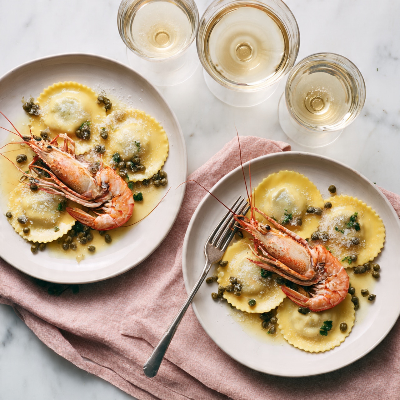 Two white plates sit on a white marbled surface with a soft pink cloth underneath. Each plate holds three round ravioli pieces arranged in a triangle, covered in a light yellow sauce with scattered green capers on top. A whole cooked langoustine with an orange shell and black eyes lies across the ravioli on each plate. Beside each plate is a silver fork resting on the edge. In the background, two clear wine glasses filled with pale golden white wine stand tall, adding elegance to the setting. photo taken with an iphone --ar 4:5 --v 7