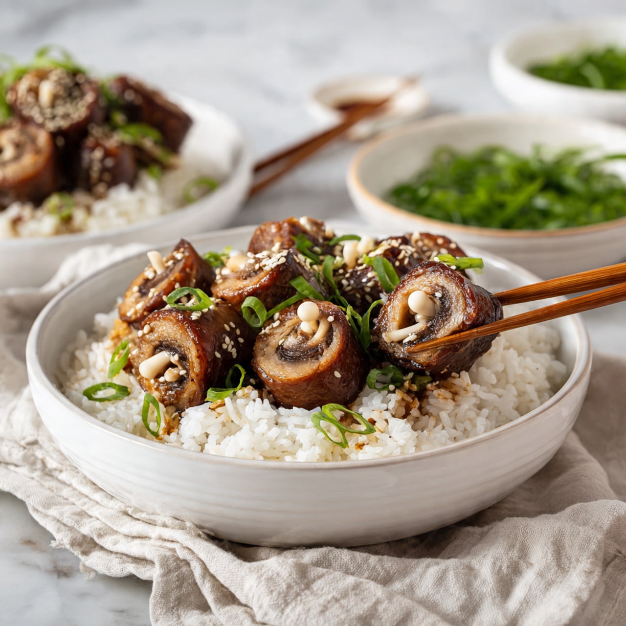 A white bowl filled with a base layer of steamed white rice, topped with several rolls of thin brown cooked meat wrapped around light beige enoki mushrooms, sprinkled with white sesame seeds and sliced green onions. A pair of wooden chopsticks held by a woman's hand picks up one of the meat and mushroom rolls above the bowl. In the background, there are more white bowls, one containing green onions, all set on a white marbled surface with a light-colored cloth underneath the main bowl. photo taken with an iphone --ar 4:5 --v 7