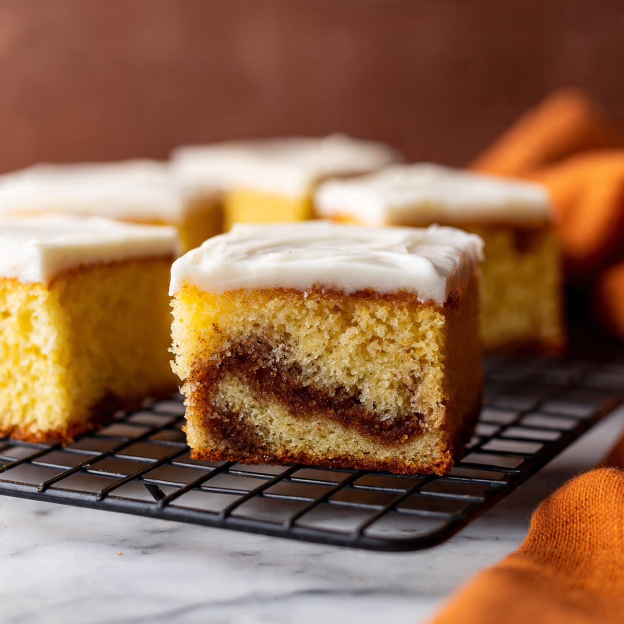 A close-up view of square-shaped cake slices showing two layers: the bottom layer is golden yellow with a moist and fluffy texture, containing a thick swirl of brown cinnamon filling near the center, while the top layer is a smooth white icing spread evenly over the cake. The cake slices sit on a black wire cooling rack, placed on a white marbled surface. There is an out-of-focus warm brown background and an orange cloth partially visible on the lower right side. photo taken with an iphone --ar 4:5 --v 7