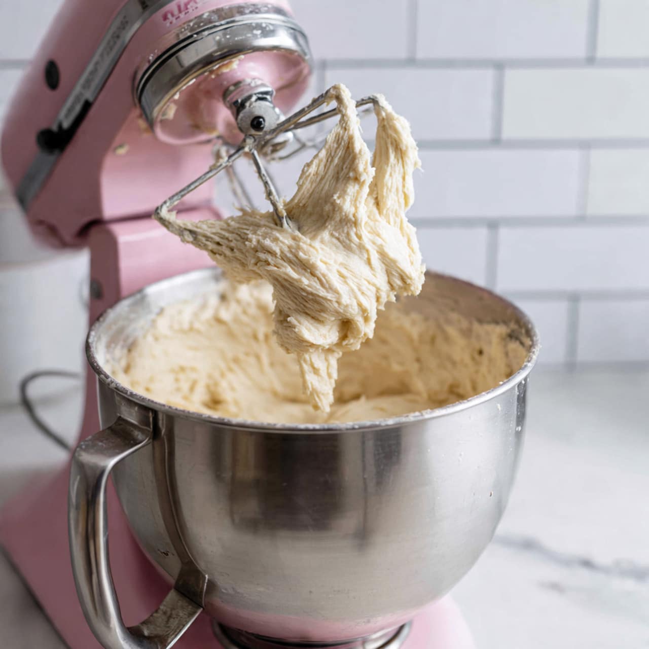 The image shows a silver metal mixing bowl attached to a pink stand mixer filled with soft, creamy, pale yellow dough. The dough has a smooth and slightly fluffy texture, spread evenly inside the bowl. A silver mixer beater with dough clinging thickly to its blades is lifted above the bowl, showing the sticky, rich consistency of the mixture. The background has white subway tiles, and the surface below is a white marbled texture. photo taken with an iphone --ar 4:5 --v 7
