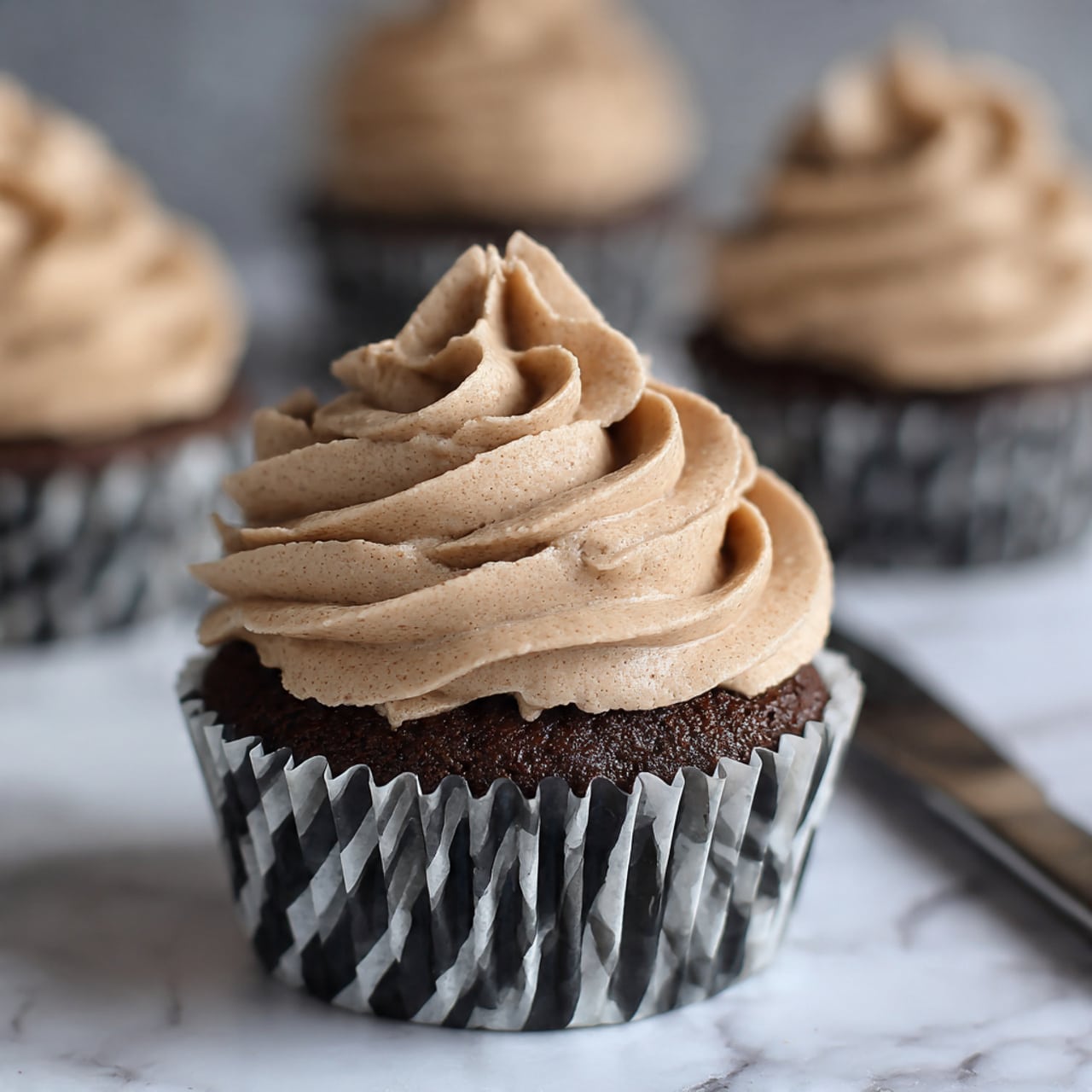 The image shows a dark chocolate cupcake with a deep rich color in a black and white striped paper liner. On top, there is one large swirl layer of light brown frosting with a smooth, creamy texture that has soft, curved ridges and peaks, creating a flower-like shape. In the background, there are blurred cupcakes with the same frosting and liners. The cupcake rests on a white marbled surface, and a silver knife is partially visible to the right side of the cupcake. photo taken with an iphone --ar 4:5 --v 7
