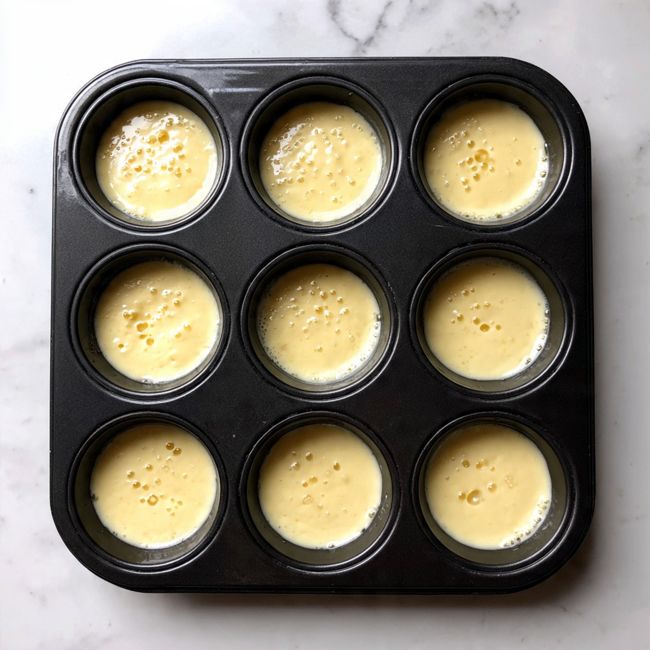 A black muffin pan with twelve cups filled with pale yellow batter that has small bubbles on its surface, showing signs of rising. The batter is mostly smooth but some cups have a few drips on the edges. The pan rests on a white marbled surface with some batter drops around it. photo taken with an iphone --ar 4:5 --v 7