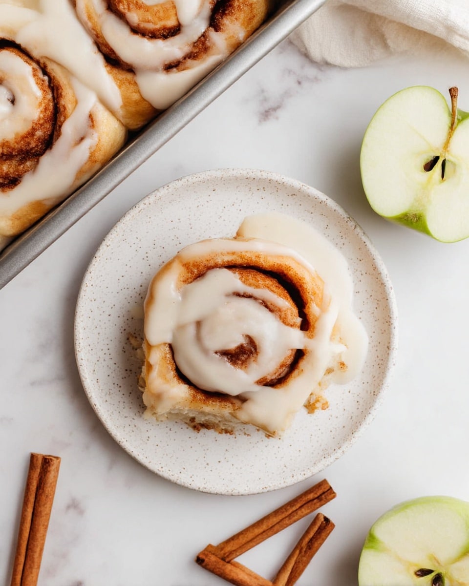 The image shows a close-up of golden brown cinnamon rolls tightly packed in a silver baking tray. The rolls have several visible layers of soft dough swirled with a slightly darker cinnamon filling, creating a spiral pattern. A white icing is being poured over the rolls from above, with some of it already settling on the top spirals, giving a smooth, creamy texture contrast to the warm baked surface. The background is a white marbled texture. photo taken with an iphone --ar 4:5 --v 7