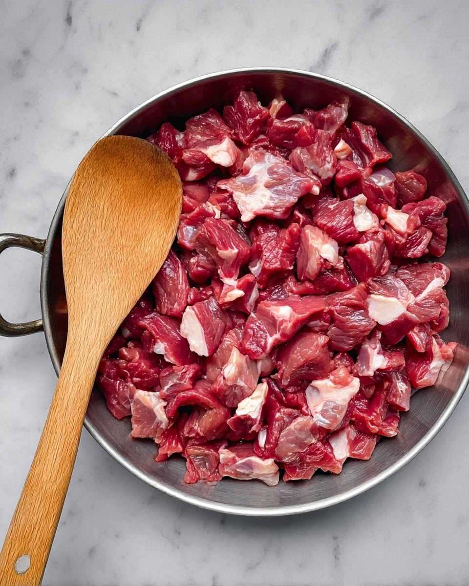 A large white metal pan with two handles holds a dish of cooked small brown meat pieces mixed with red bits, soaked in an oily brown sauce. The edges of the pan have brown splatters from the sauce. Two pieces of light beige flatbread rest on the left side inside the pan. To the right of the pan, on a white marbled surface, there is a small glass bowl with green seeds and a small white bowl with diced red tomatoes and green cucumbers. A folded pink cloth is partially visible in the top left corner. Photo taken with an iphone --ar 4:5 --v 7