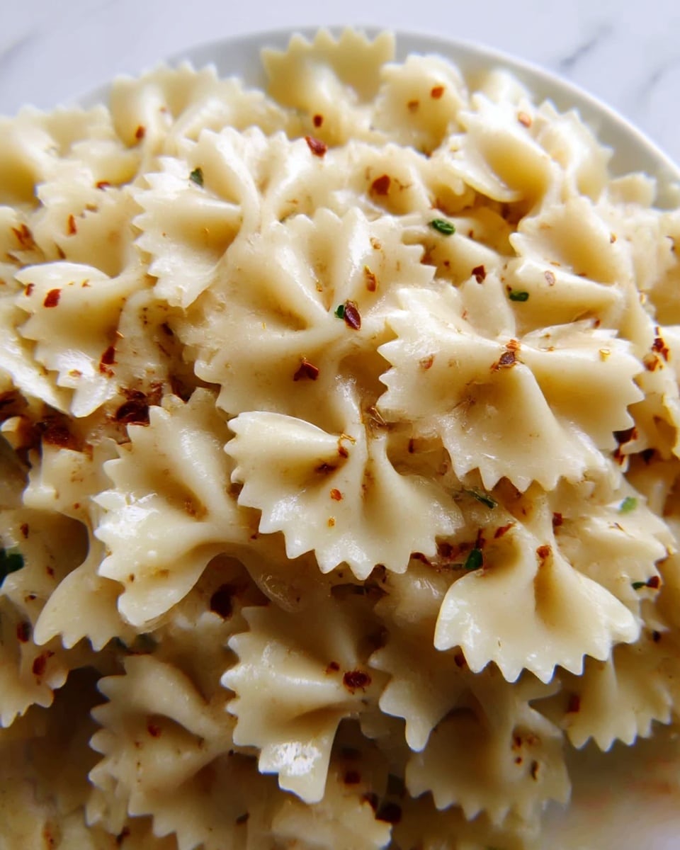 The image shows a close-up of a white plate filled with cooked farfalle pasta. The pasta is light cream in color with a soft, slightly glossy texture. Small bits of seasoning, including green herbs and red chili flakes, are lightly scattered across the top layer of the pasta, giving it a speckled look. The white plate sits on a white marbled surface, and the overall scene is bright and simple. photo taken with an iphone --ar 4:5 --v 7