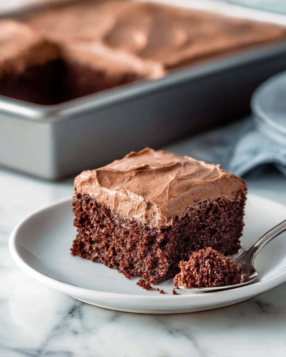 A square piece of dark brown chocolate cake with a thick, smooth layer of light brown chocolate frosting on top. The cake looks moist and soft with a crumbly texture inside. A small piece of cake is on a spoon beside the cake on the white plate, showing the rich texture clearly. The background shows the cake pan with the remaining cake, all on a white marbled surface. Photo taken with an iphone --ar 4:5 --v 7