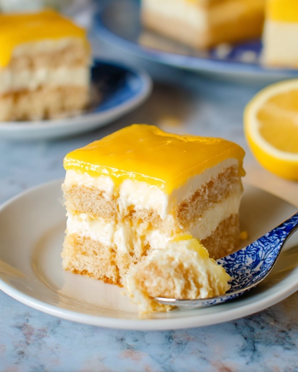 The image shows a square slice of layered lemon dessert on a white plate with a blue and white patterned spoon next to it. The dessert has three layers: the bottom and middle layers are light tan cake pieces with a soft spongy texture, separated by a thick creamy white layer in the middle. The top layer is a smooth, shiny bright yellow lemon glaze. A bite-sized piece of the dessert on the spoon shows the creamy and cake layers clearly. In the background, there is a second slice of the same dessert on another white plate and a lemon wedge is partly visible. The surface is a white marbled texture. photo taken with an iphone --ar 4:5 --v 7
