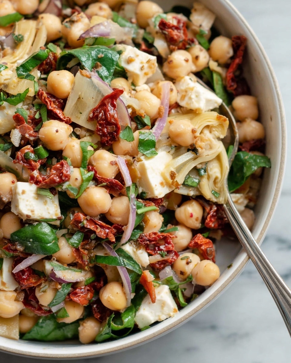 A close-up view of a white bowl filled with a colorful bean salad with several layers visible; the bottom layer is light beige white beans mixed with round, pale yellow chickpeas, above that are soft artichoke heart pieces in pale yellow with a smooth texture, scattered with deep red sun-dried tomatoes that have a wrinkled appearance, and small cubes of white cheese mixed evenly throughout. Fresh green spinach leaves and chopped green herbs sit on top, adding vibrant color, while thinly sliced red onions add a touch of purple. A silver spoon is partially visible inside the bowl, and the whole scene rests on a white marbled surface. photo taken with an iphone --ar 4:5 --v 7