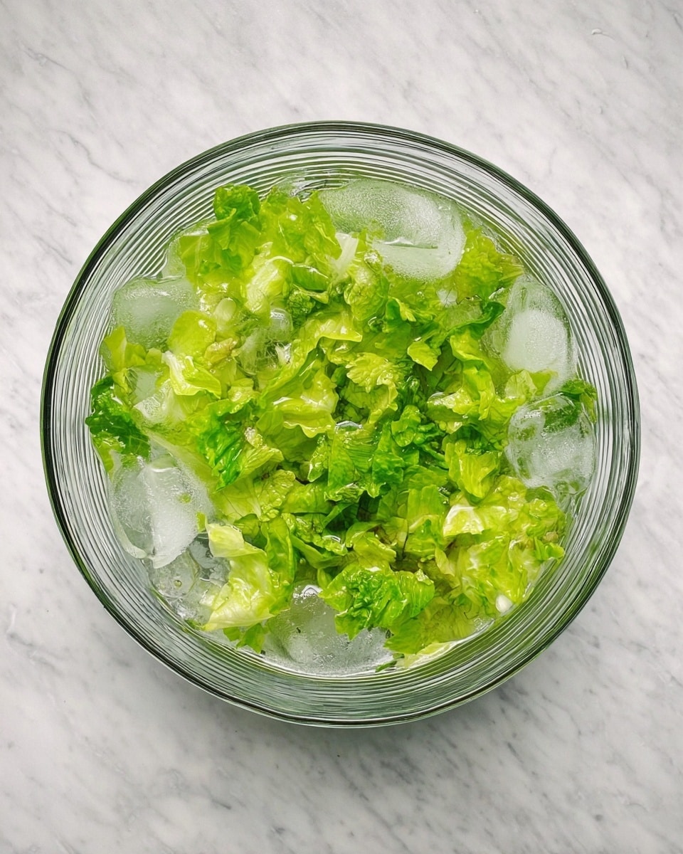 A clear round glass bowl filled with crushed ice at the bottom, layered with bright green torn lettuce leaves floating on top in water. The texture of the lettuce is fresh and slightly crinkled, with light and dark green shades mixed. The bowl sits on a white marbled surface. photo taken with an iphone --ar 4:5 --v 7