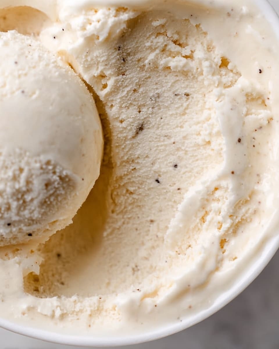 A close-up image of creamy vanilla ice cream with visible vanilla bean specks. The ice cream shows a smooth and soft texture with some areas scooped out, revealing a slightly fluffy and aerated inside. The ice cream fills a white bowl, with the curved edges visible around it. The background and surface are a white marbled texture, making the creamy light beige color of the ice cream stand out. Photo taken with an iphone --ar 4:5 --v 7