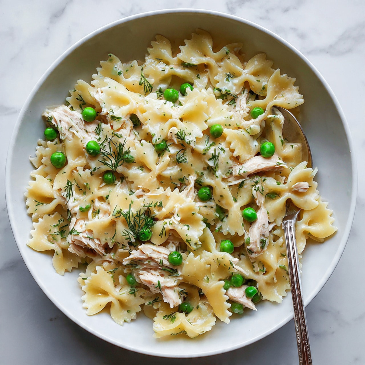 The image shows a close-up of a white bowl filled with creamy pasta made of farfalle shapes, pale yellow in color with a smooth texture. Mixed inside are light beige pieces of shredded chicken and bright green peas scattered evenly throughout. Small bits of fresh dill add a touch of green and fine texture across the pasta surface. A silver spoon rests inside the bowl on the right side, holding a small serving of the pasta, peas, and chicken. The background has a white marbled texture, creating a clean and simple look. photo taken with an iphone --ar 4:5 --v 7