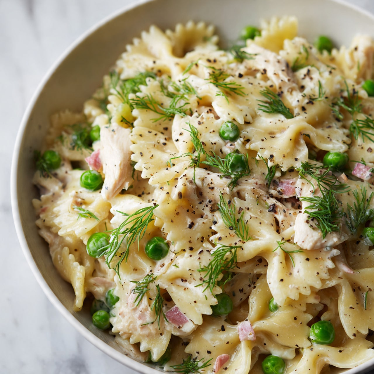 A close-up view of a white bowl filled with creamy bowtie pasta. The pasta is pale yellow and mixed with small pieces of light pink chicken, bright green peas, and bits of cream-colored diced vegetables. Fresh green dill sprigs are scattered on top, along with a light sprinkling of black pepper. The sauce coats the pasta softly, giving it a smooth texture. The bowl rests on a white marbled surface. photo taken with an iphone --ar 4:5 --v 7