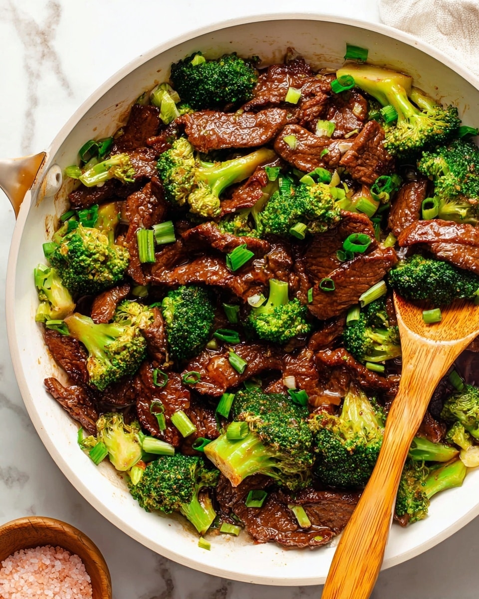 The image shows a white pan full of beef and broccoli stir-fry. The dish has two main layers: the first layer consists of thin, dark brown beef slices with a shiny sauce covering them, and the second layer features bright green broccoli florets and chopped green onions scattered evenly on top and mixed throughout. A wooden spoon is resting inside the pan, slightly lifting some beef and broccoli. The pan sits on a white marbled surface with a small wooden bowl of pink salt nearby. Photo taken with an iphone --ar 4:5 --v 7