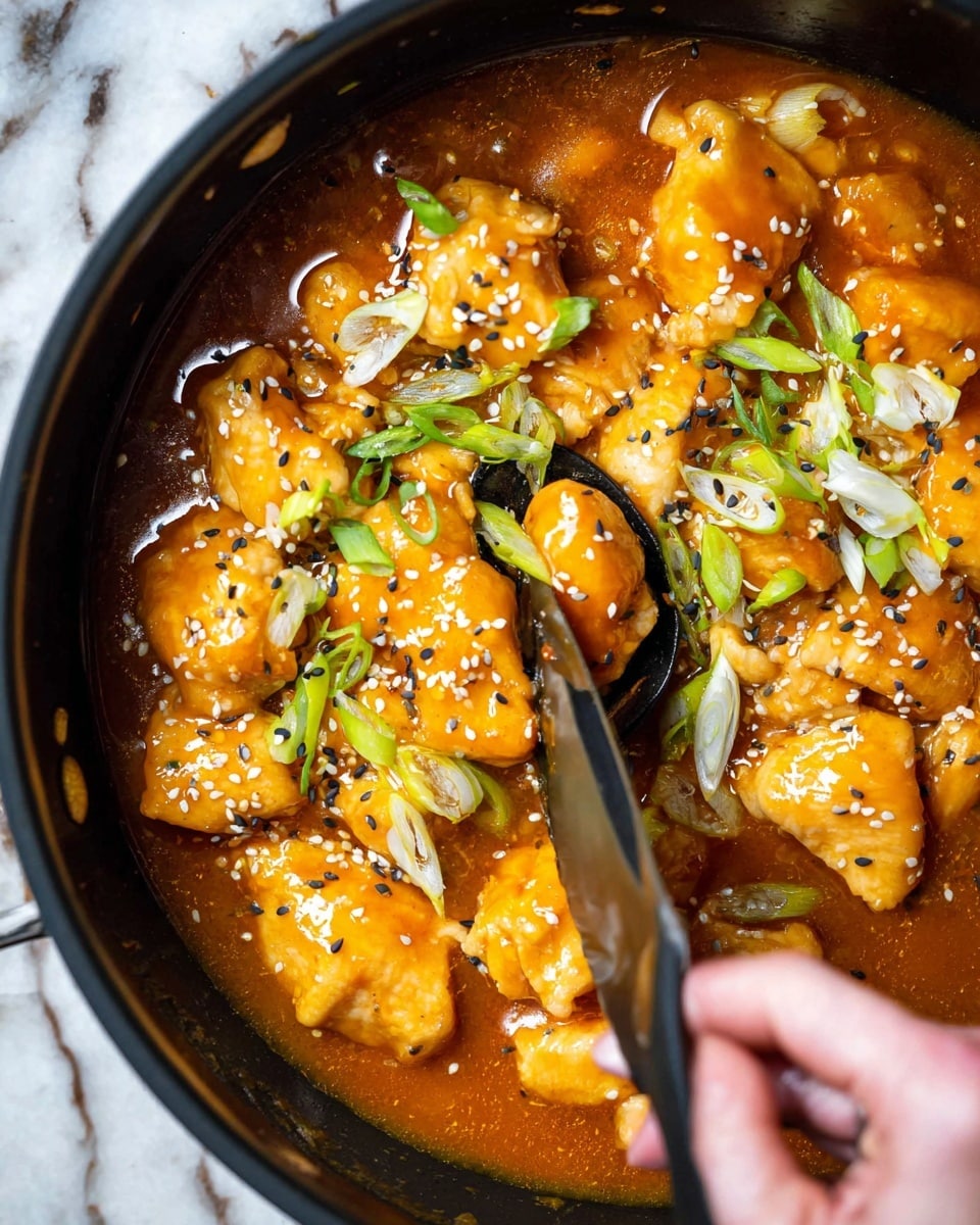 A close-up view of a black pan filled with golden brown pieces of cooked chicken in a thick, bright orange sauce. The chicken pieces are topped with white and black sesame seeds and sliced green onions. A woman's hand holds a black spoon stirring the chicken, while a silver knife lifts a piece of chicken covered in the rich sauce. The background surface is a white marbled texture. photo taken with an iphone --ar 4:5 --v 7