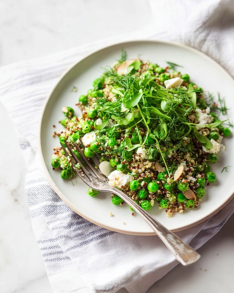 A white plate on a white marbled surface holds a fresh quinoa salad with bright green peas mixed evenly throughout. The quinoa layer is light and fluffy, speckled with small white grains. Scattered on top are thin slices of almonds and sprigs of fresh green herbs including dill and microgreens, adding texture and height. Crumbled white cheese is sprinkled lightly over the salad, contrasting with the green and beige colors. A shiny metal fork rests on the right side of the plate, partially inside the salad. The background shows a soft, white cloth with gray stripes. Photo taken with an iphone --ar 4:5 --v 7