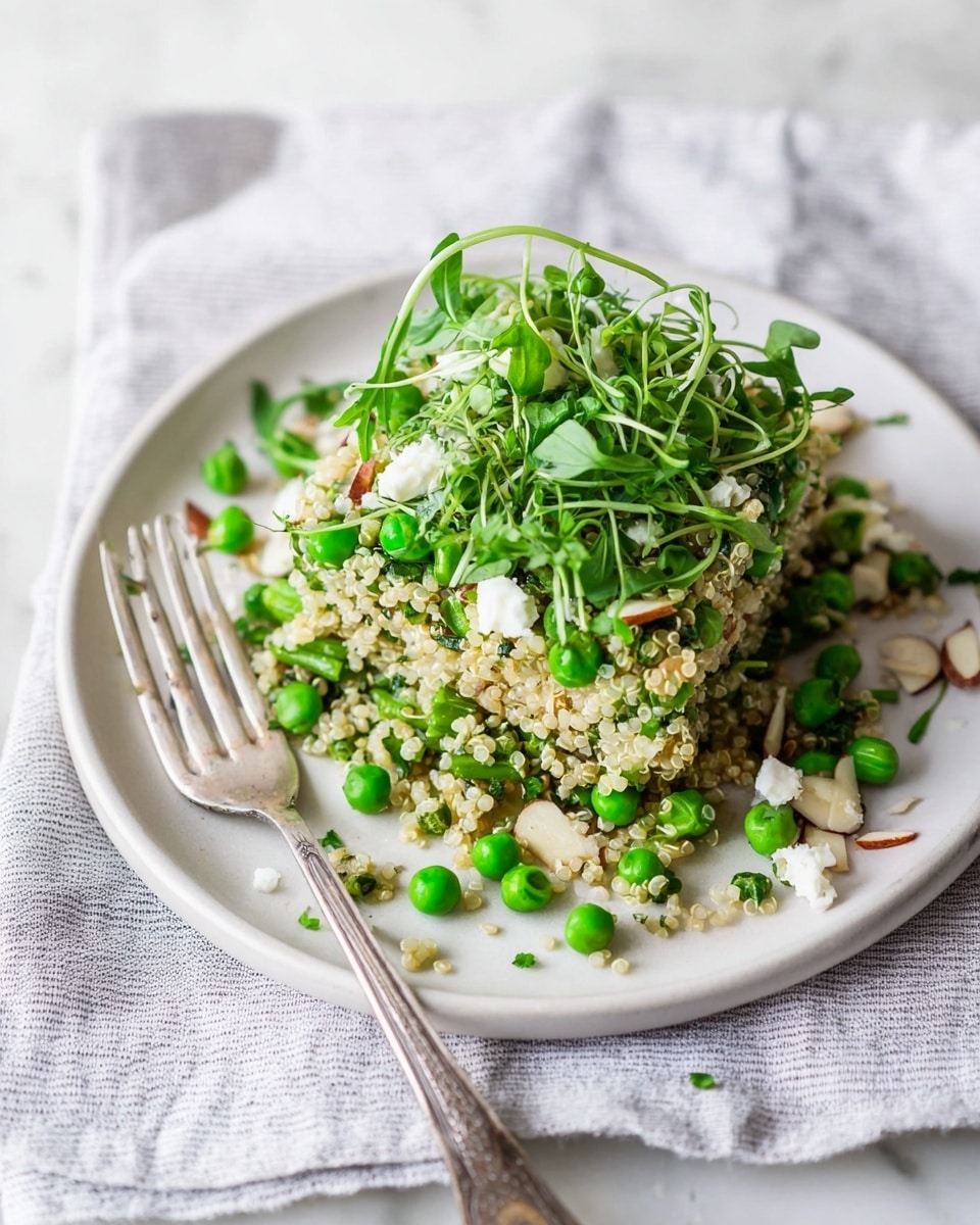 A white plate holds a fresh quinoa salad layered with bright green peas and chopped green vegetables mixed in, showing a fluffy texture beneath. On top, thin pea shoots, fresh dill sprigs, and scattered small white cheese crumbles add light color and texture. Thin pale almond slices are spread around the base, adding a crunchy look. A fork rests on the right edge of the plate. The plate is placed on a white marbled surface covered by a striped cloth with light purple and white lines. photo taken with an iphone --ar 4:5 --v 7