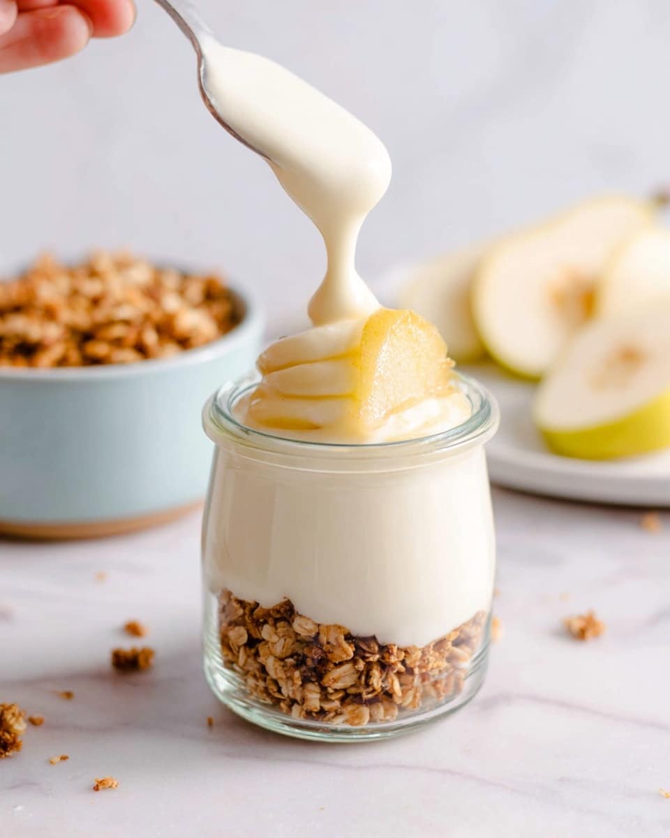 This image shows a small clear glass jar with two main layers. The bottom layer is a light brown, crumbly granola layer that fills about one-third of the jar. Above this, there is a thick, creamy white yogurt layer that fills the rest of the jar, leaving a smooth, slightly bubbly surface on top. On top of the yogurt sits a small, pale yellow curved slice, likely a piece of fruit, and a small sprinkling of granola crumbs. The jar sits on a white marbled surface, and there is a wooden spoon nearby. In the background, there is a soft-focus second jar with the same contents and a pale blue bowl filled with granola. Photo taken with an iphone --ar 4:5 --v 7