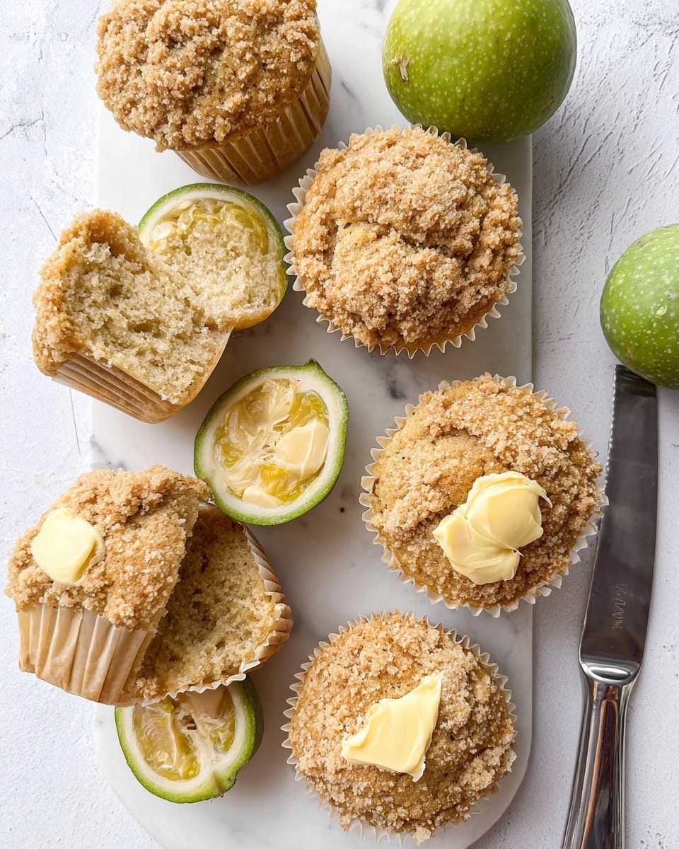 The image shows a white rectangular plate with four golden-brown muffins topped with oat crumb, arranged on the left side. One muffin near the top right is split in half, revealing a soft, light brown inside with two small dollops of yellow butter on each half. Next to the halved muffin, there are two whole bright green feijoa fruits and one feijoa cut in half, showing its pale green inside with seed patterns. To the right side of the plate, there is a silver butter knife resting on the white marbled surface. The scene is bright with natural light, creating soft shadows. photo taken with an iphone --ar 4:5 --v 7