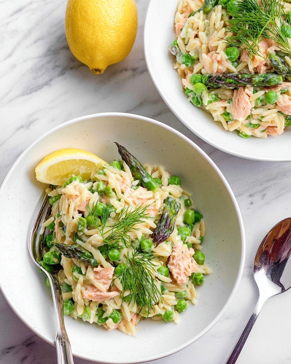 Two white bowls filled with a creamy rice dish that has layers of light beige rice mixed with green peas and asparagus pieces, along with small chunks of orange salmon scattered throughout. Each bowl is garnished with fresh green dill on top, and a bright yellow lemon wedge is placed on the edge of the closest bowl. One bowl is fully visible while the other is partially shown with a silver spoon resting inside. A silver fork lies on the white marbled surface near the bowls. The overall look is fresh and colorful with a smooth creamy texture. photo taken with an iphone --ar 4:5 --v 7