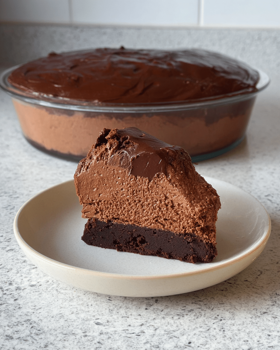 A slice of chocolate mousse cake with two layers sits on a simple white plate; the bottom layer is a dark chocolate cake with a light and airy texture, and the top layer is a thick, creamy chocolate mousse with a rich, smooth look. Behind the plate, there is a clear round glass dish filled with the same chocolate mousse cake, showing a glossy, thick chocolate glaze covering the top, with a chunk missing on one side. The surface underneath both the plate and glass dish is a white marbled texture. photo taken with an iphone --ar 4:5 --v 7