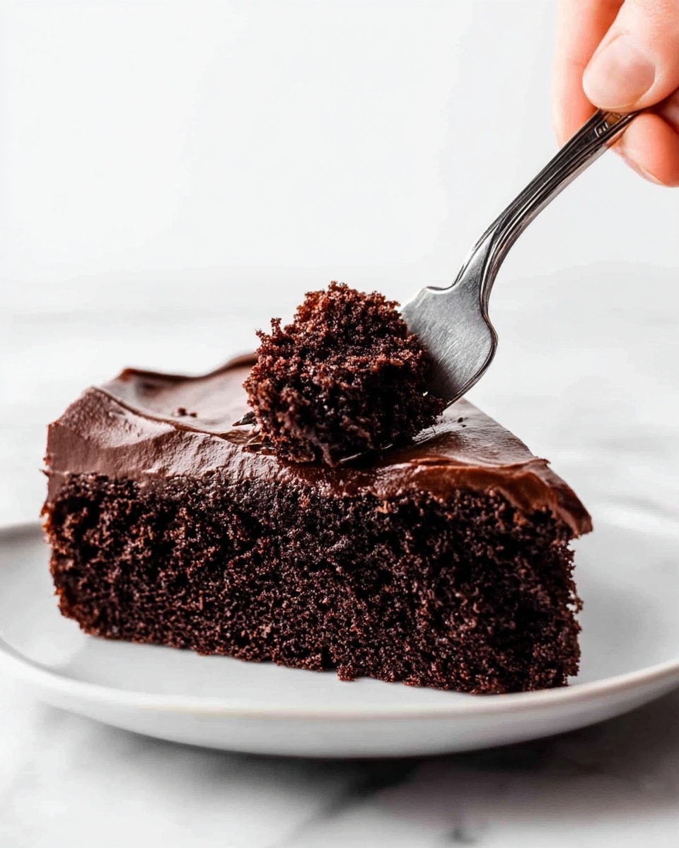 A thick, dark brown chocolate cake slice sits on a white plate, showing a soft and moist texture with tiny crumb details. The top layer is covered with smooth, glossy chocolate frosting that has a rich, creamy look and slightly drips over the edge. A silver fork rests beside the cake slice on the plate. The setting has a white marbled surface in the background. photo taken with an iphone --ar 4:5 --v 7