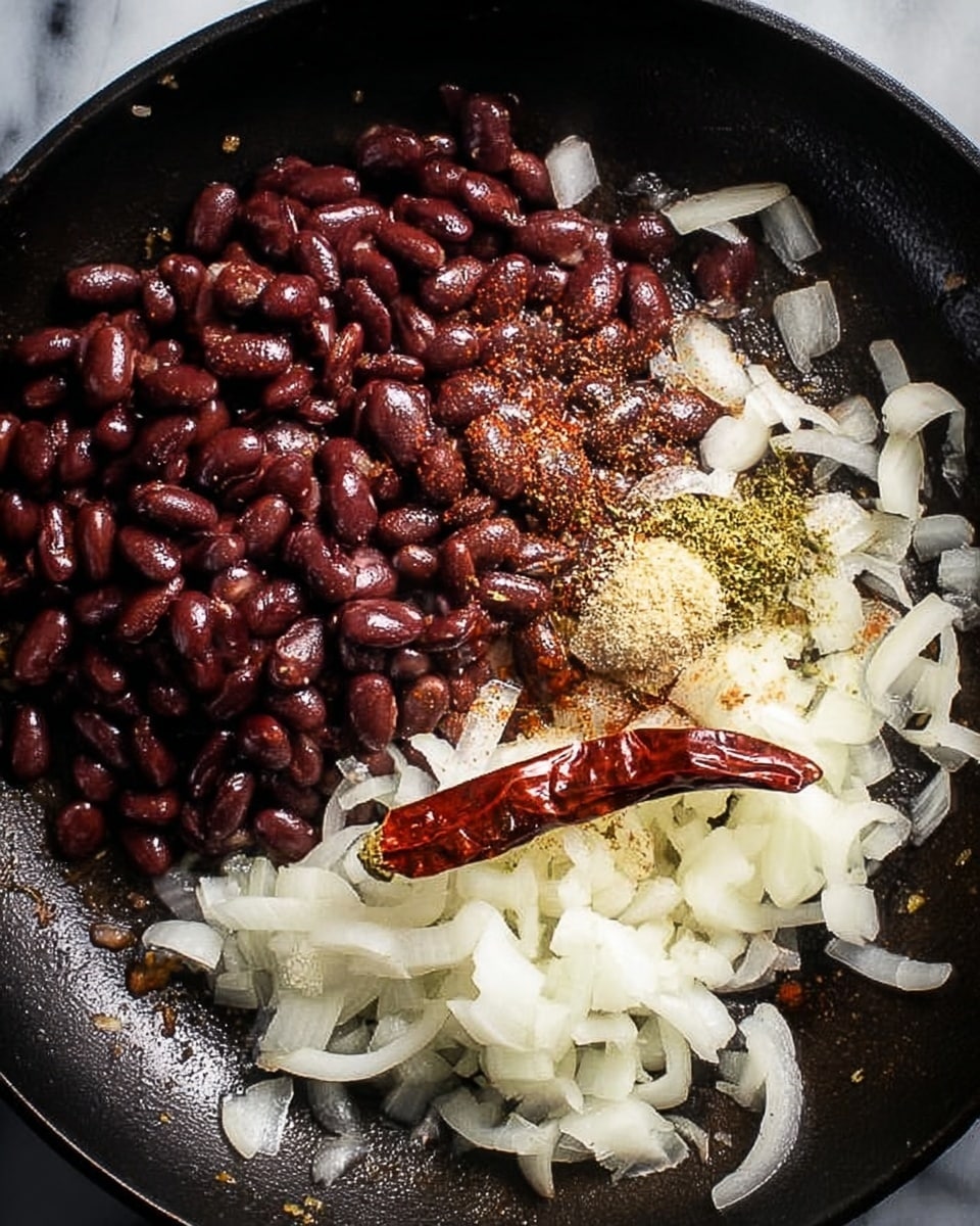 The image shows a dark skillet filled with a mix of ingredients. On the left side, there is a mound of shiny, dark red beans. On the right side, sliced white onions are spread out, some translucent as if lightly cooked. On top of the onions and beans, there is a sprinkle of light-colored spices and herbs, adding texture and contrast. Near the bottom right, a single large dried red chili rests on the onions. The skillet's surface is slightly oily, giving a subtle shine to the ingredients. The background is a white marbled texture photo taken with an iphone --ar 4:5 --v 7