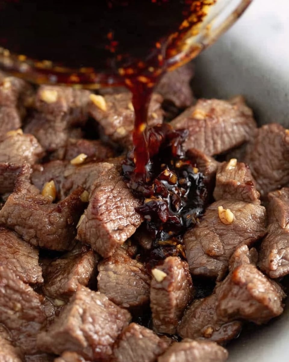A close-up shows many small pieces of cooked beef with a slightly brown and gray color, resting in a pan. A dark, glossy sauce is being poured over the beef from above, with some sauce droplets visible mid-pour. The texture of the beef looks tender with some small bits of garlic or seasoning on top. The background is a white marbled surface. Photo taken with an iphone --ar 4:5 --v 7