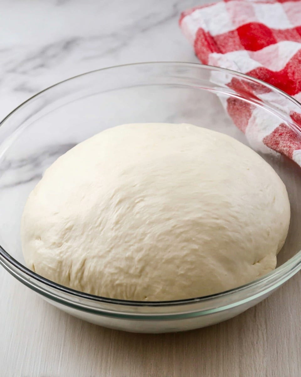 A clear glass bowl holds one large, smooth, round ball of pale white dough, showing a soft and slightly shiny texture. The dough sits flat in the bowl with faint wrinkles and a few small dimples on its surface. The bowl is placed on a white marbled surface, with part of a white and red checkered cloth visible in the background. Photo taken with an iphone --ar 4:5 --v 7
